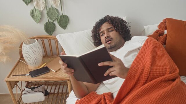 Young indian man reading book on bed in studio bedroom, wrapped in orange blanket with rattan nightstand and lamp visible; cozy comfort serenity.