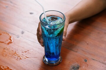 Close up of hand holding a fresh blue mojito drink on wooden table with natural lighting. © ROHE Creative Studio