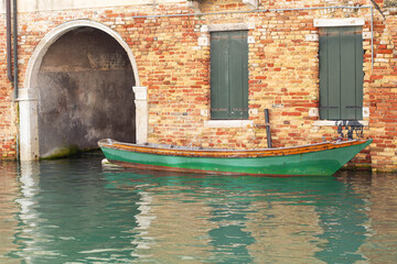 Green wooden boat moored beside a weathered brick wall with arched entrance and green shutters reflecting in the calm water of a canal in Venice, Italy