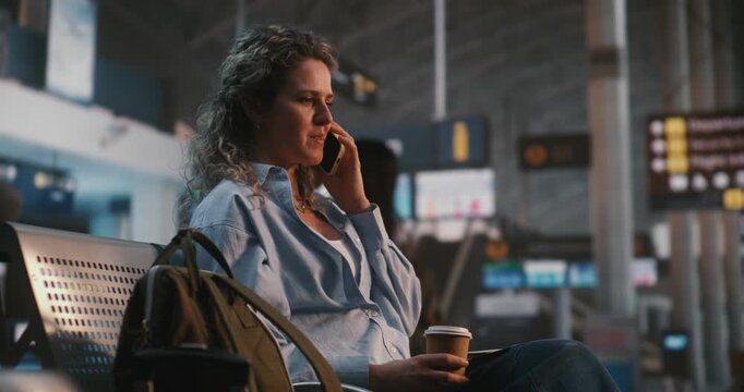 Young Woman Sits Comfortably In Airport Lounge, Enjoying Coffee While Having Cheerful Phone Conversation. Stress Free Travel Experience, Positive Emotions, and Staying Connected With Loved Ones.