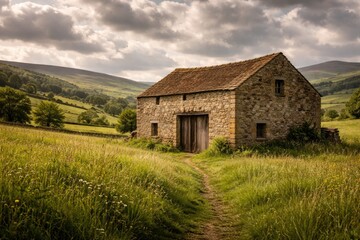 Obraz premium Old stone barn standing in a lively grassy meadow with rolling hills and cloudy sky