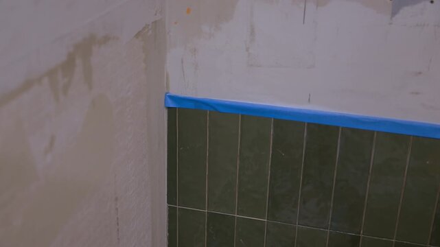 Bathroom renovation in progress with partially tiled wall and exposed plaster surface. Blue painters tape marks alignment edge for tile installation. Interior construction.