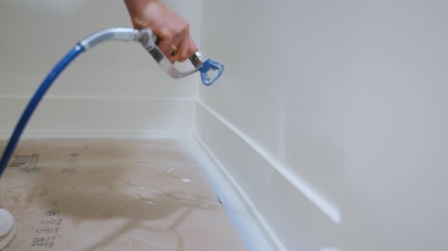Worker using a spray gun to paint an interior wall during renovation. Professional painting process with airless sprayer, 