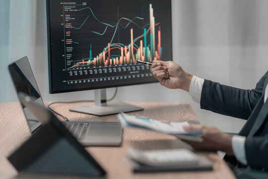 A business person is analyzing data on a computer screen. The screen displays colorful line graphs and bar charts, or performance indicators. The person is holding a pen and pointing at the chart.