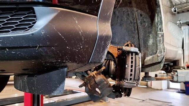 Front end of vehicle lifted in workshop with wheel removed, exposing brake caliper, rotor, and suspension components, highlighting automotive repair, inspection, and maintenance process