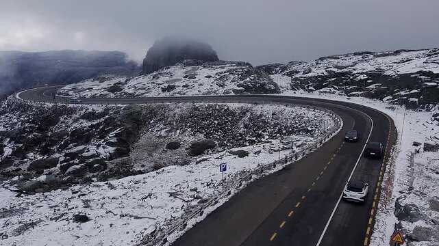 Aerial tracking cars driving winding road N339 through snow covered Serra da Estrela mountains, Portugal, with Cantaro Magro peak in winter
