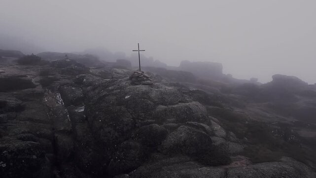 Summit cross stands on rugged granite rocks in Serra da Estrela Portugal surrounded by dense fog and harsh mountain terrain creating moody atmospheric scene, aerial drone slow glide shot