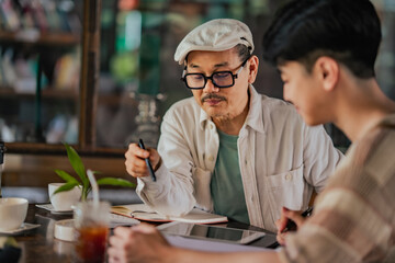 Fototapeta premium Asian father teaches Gen Z son cafe startup accounting at small family business table. SME planning, expense tracking, digital finance learning in cozy coffee shop environment with tablet and notes.