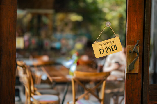 Closed sign hanging on cafe door with green plants in foreground and blurred interior. Represents business closed status, quiet atmosphere, small business concept and hospitality downtime.