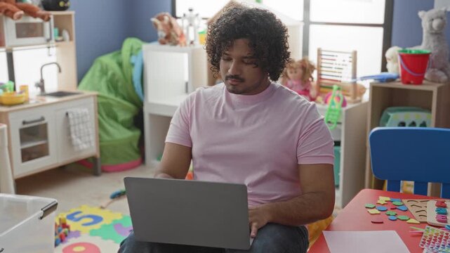 Man typing on laptop in a kindergarten playroom building surrounded by foam mats, toys and a doll at a small table; quiet concentration.