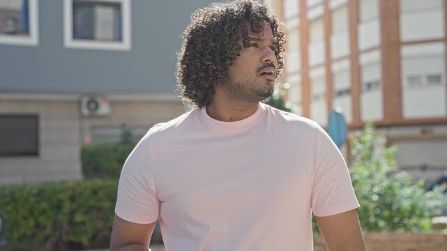 Man in pink tshirt holding smartphone with both hands, scrolling and looking down amid residential buildings and sidewalk on a street; concentration.