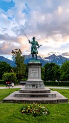 Obraz premium A classical statue of a man holding objects stands in a park with mountains in the background under a cloudy sky