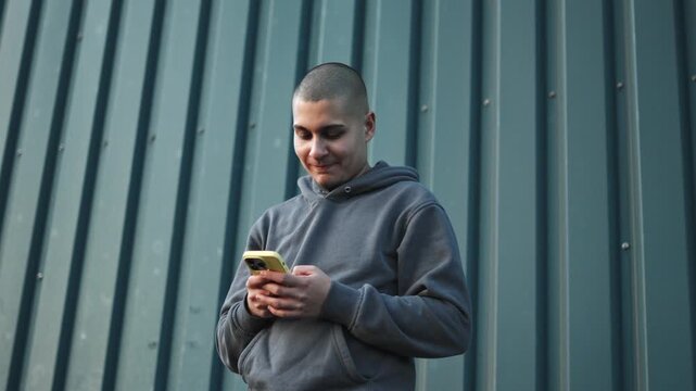 Young man with buzz cut smiling while texting on smartphone against a corrugated metal wall