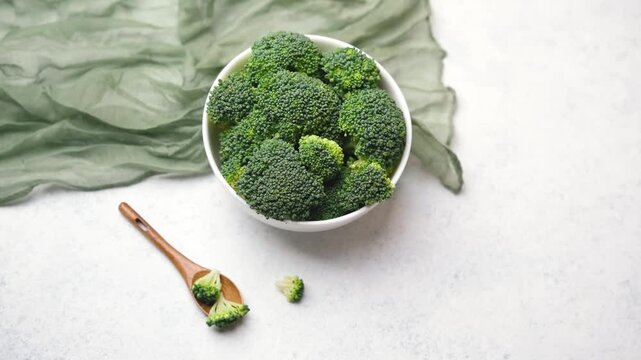 Fresh green broccoli florets in white bowl with wooden spoon on textured surface