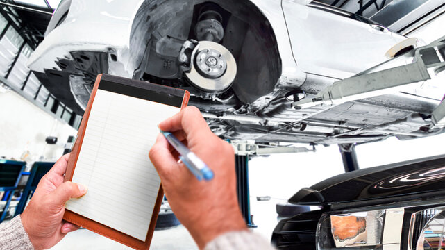 Car Inspection Checklist, A mechanic writes on a blank notepad while performing a vehicle inspection on a car raised on a hydraulic lift in a modern auto garage.