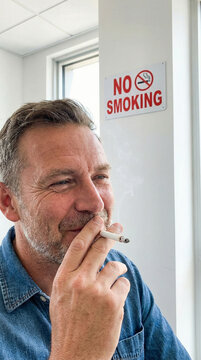 Smiling Man Defying Rules by Smoking Near No Smoking Sign