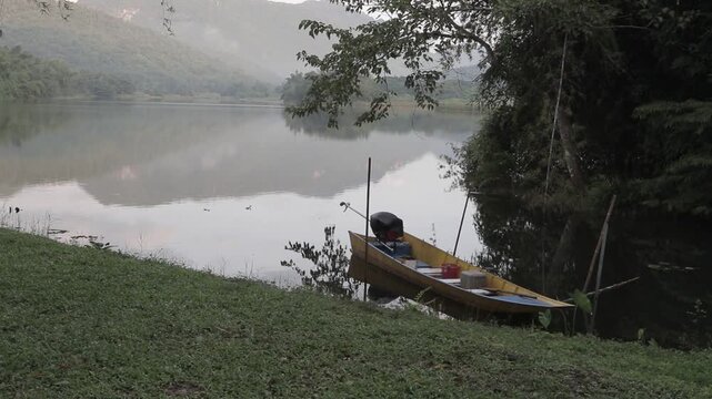 A traditional long-tail boat moored by a calm river with mountain reflections in Kanchanaburi, Thailand. Tranquil riverside lifestyle scenery.