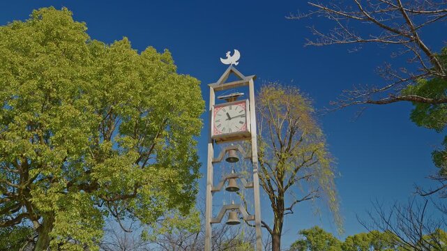 A tall wooden clock tower featuring three bells and a bird ornament at the top, standing among green trees in Gyosen Park on a clear day.