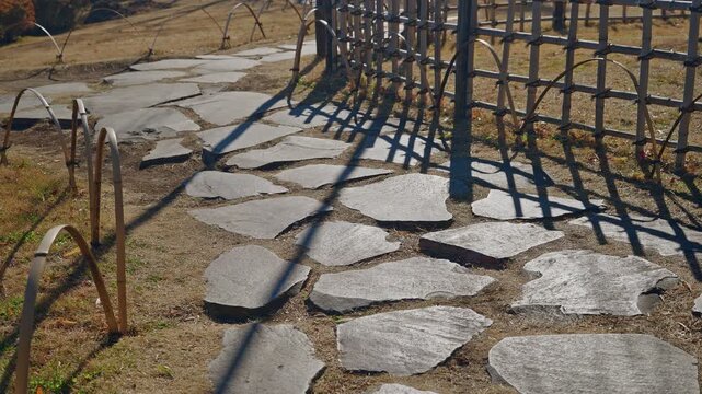 A slow tilt up capturing the texture of an irregular stone path (tobi-ishi) and a low bamboo fence with strong shadows in a traditional Japanese garden.