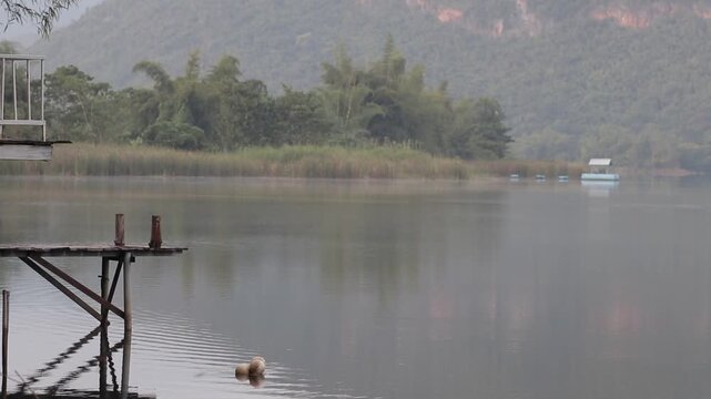Wooden pier over a calm, misty river with mountains in the background at Kanchanaburi, Thailand. A very quiet and tranquil morning scene.