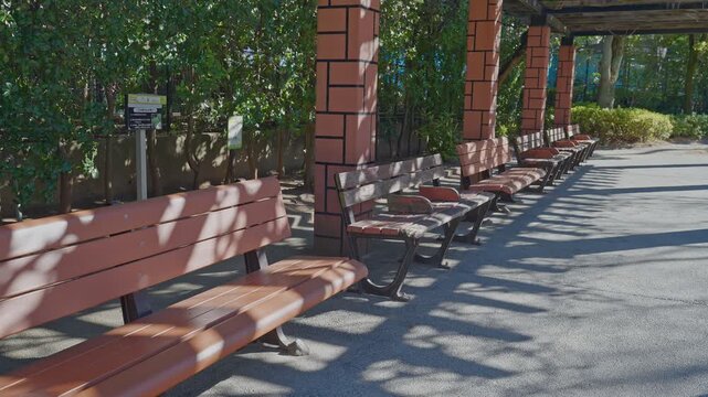 A row of wooden benches under a brick-pillared pergola in a quiet park, featuring beautiful dappled sunlight and shadows on a bright day.