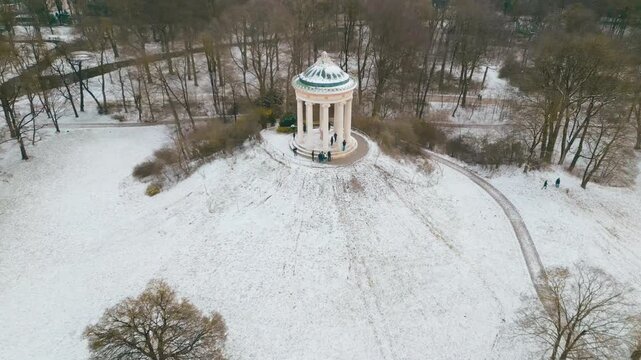 Drone panning away from the Monopteros in Munich&rsquo;s English Garden, surrounded by snow on a sunny winter morning with serene park atmosphere.