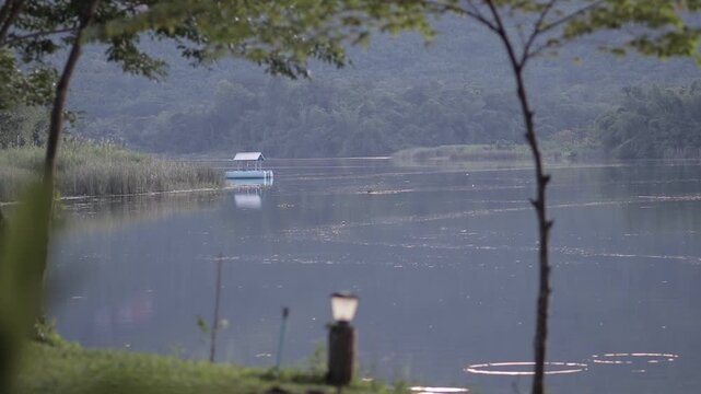 A peaceful view of a floating house on a calm river seen through trees in Kanchanaburi, Thailand. A serene and private tropical morning scenery.