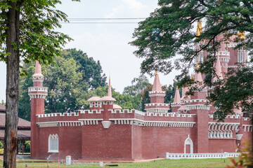 Red brick castle with pointed turrets and white trim. Green trees frame the structure on either side. Bright sky above with light cloud cover. Castle stands on grassy grounds with low fence © jinhongljh