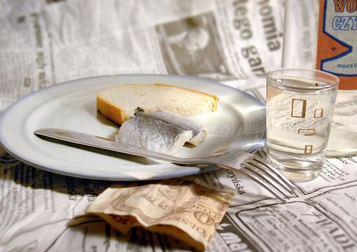A picture in the style of the Polish People's Republic (PRL). On a newspaper, as if on a tablecloth, a plate with a herring and a piece of bread. Next to it, a glass of vodka. 
