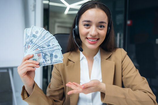 Webcam view of Emotional caucasian businesswoman in wireless headset explaining ways of getting great income holding bunch of dollars