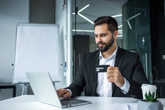 bearded businessman doing online shopping and bank money transfer, freelancer holding bank credit card and smartphone, man sitting at desk inside office.