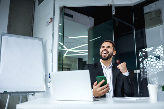 Happy millennial guy feel overjoyed read good news in email. Win. Portrait of excited mature man celebrating success shaking clenched fist using cellphone, sitting at desk with laptop, office interior