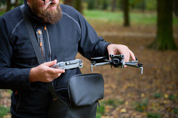 A man with a beard stands in a peaceful park, getting ready to launch his drone. The colorful...