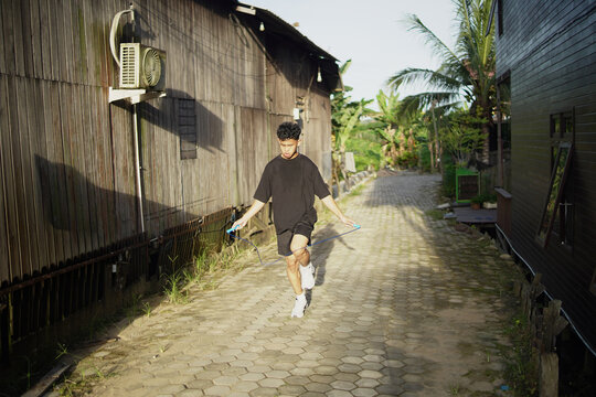 Young Asian man skipping rope outdoors for fitness. Focused athletic male exercising cardio workout in home background, daylight