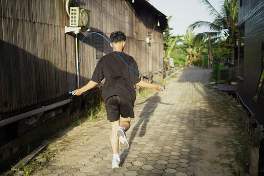 Young Asian man skipping rope outdoors for fitness. Focused athletic male exercising cardio workout in home background, daylight