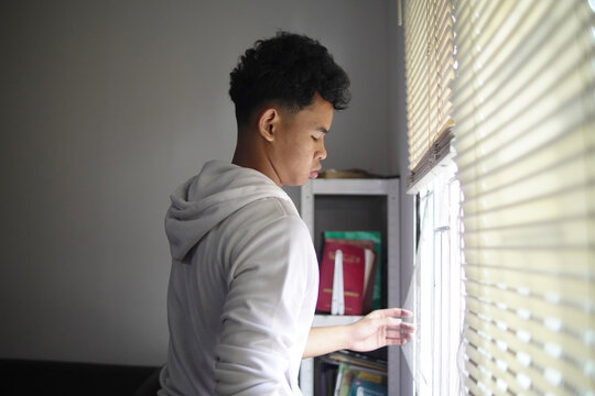 Young Asian man opening or closing window blinds at home, looking out for hope or privacy, morning routine or mental health concept