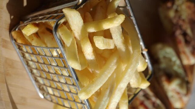 Crispy golden french fries in metal basket moving slowly over wooden board with sandwiches.