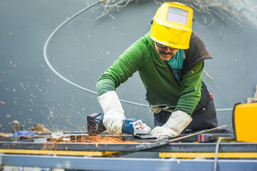 A worker is cutting metal in a workshop. The sparks fly as the tool grinds against the metal surface. The worker is wearing a safety helmet and gloves