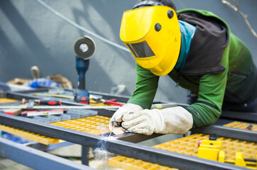 A worker person is working in a workshop with tools and metal. They wear safety gear and focus on shaping a piece of metal. The workspace is organized with various tools around
