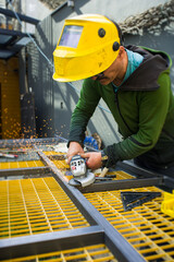 A worker cuts metal with a tool on a construction site. Sparks fly as he focuses on his task. The bright yellow grid beneath him adds color to the scene
