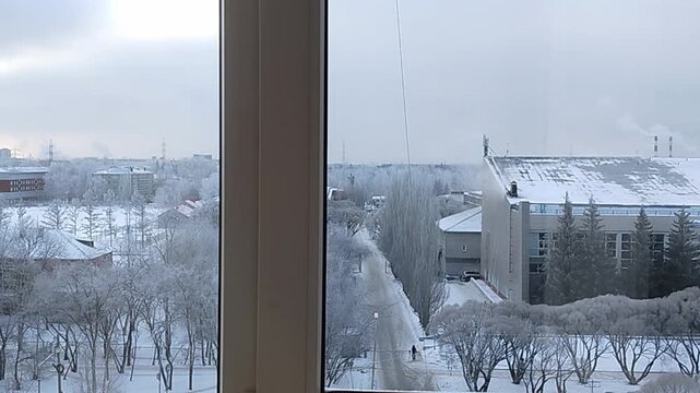 Winter landscape view from a window showcasing snow-covered trees, buildings, and a pathway, capturing the serene atmosphere of a cold day in a city