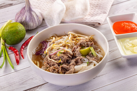 Traditional Vietnamese Pho Bo soup with sliced beef, rice noodles, and fresh herbs on a white wooden table.