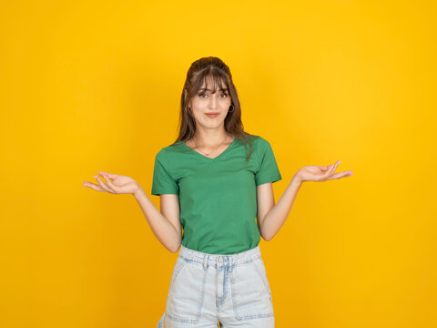 Confused caucasian woman shrugging with both hands, expressing uncertainty or indecision concept idea, wearing green t shirt and denim, isolated on bright yellow studio background with copy space.