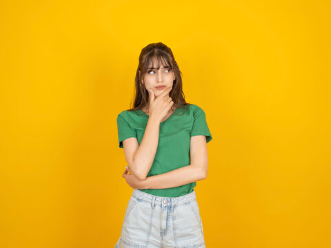 Thoughtful caucasian woman touching chin and looking aside, considering a decision or idea concept, wearing green t shirt and denim, isolated on bright yellow studio background with copy space.