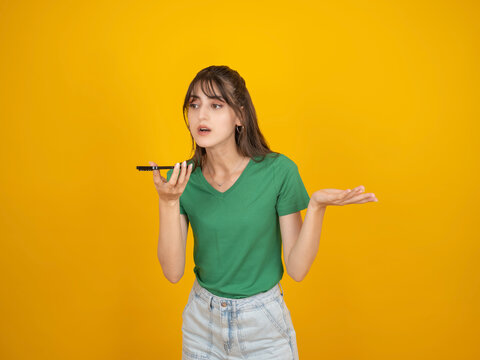 Confused caucasian woman holding smartphone and speaking with questioning expression, gesturing with open hand, wearing green t shirt and denim, isolated yellow studio background with copy space.