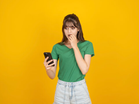 Worried caucasian woman looking at smartphone while biting nails, showing anxiety and nervous reaction concept, wearing green t shirt and denim, isolated yellow studio background with copy space.