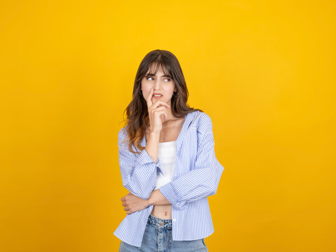 Caucasian woman thinking with doubtful expression, touching lips while looking aside, considering a decision, wearing striped shirt and denim, isolated yellow studio background with copy space.