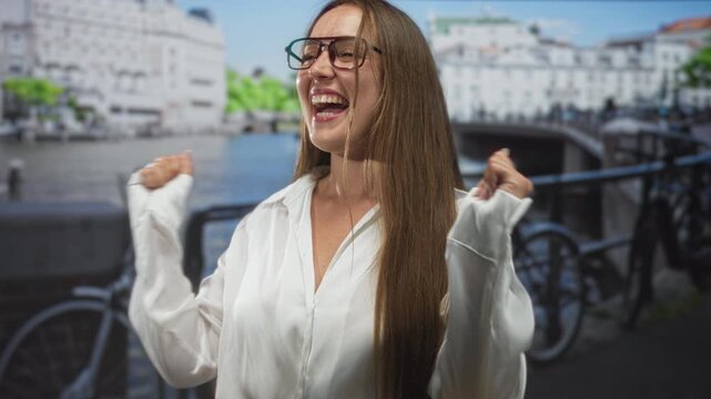 Woman clapping hands on canal street in amsterdam, smiling with glasses and long hair, white shirt; carefree joy.