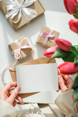Fototapeta premium Vertical shot of woman's hands holding a blank white card over table with red tulips and craft gifts. Greeting card mockup