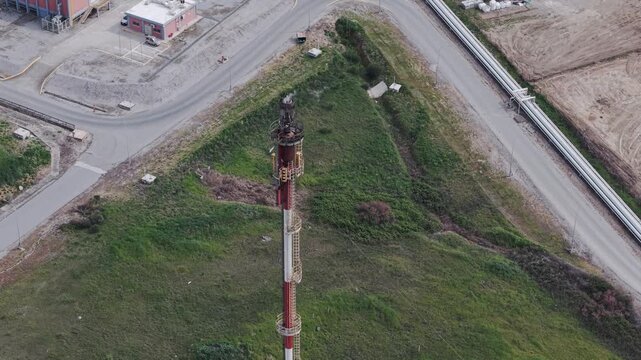 Aerial view of an industrial area featuring a tall tower with a flame contrasting against the green grass, Thessaloniki, Thessaloniki, Greece.